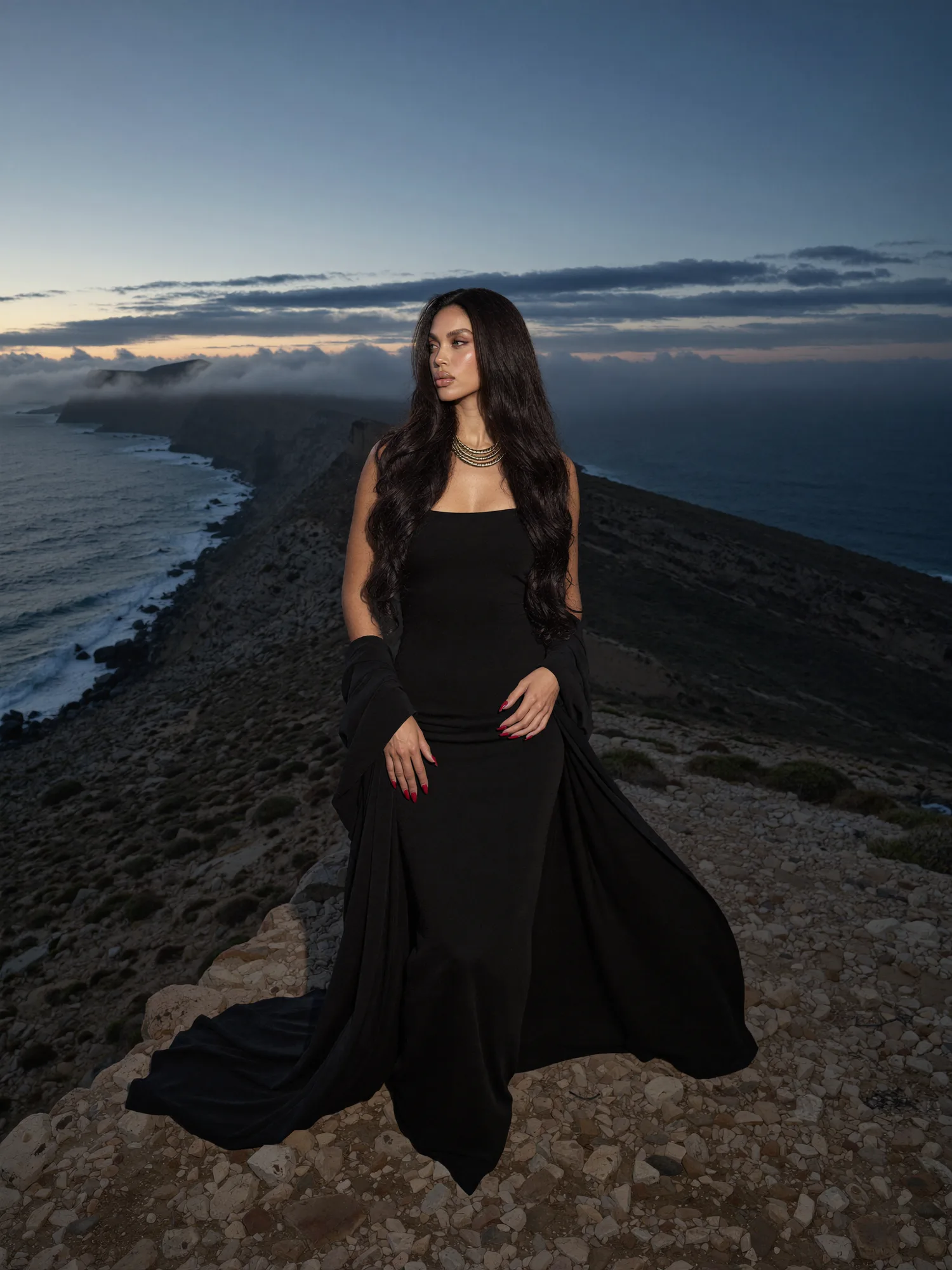 Seven Yuno, the founder, in a long black gown on a coastal ridge at dusk
